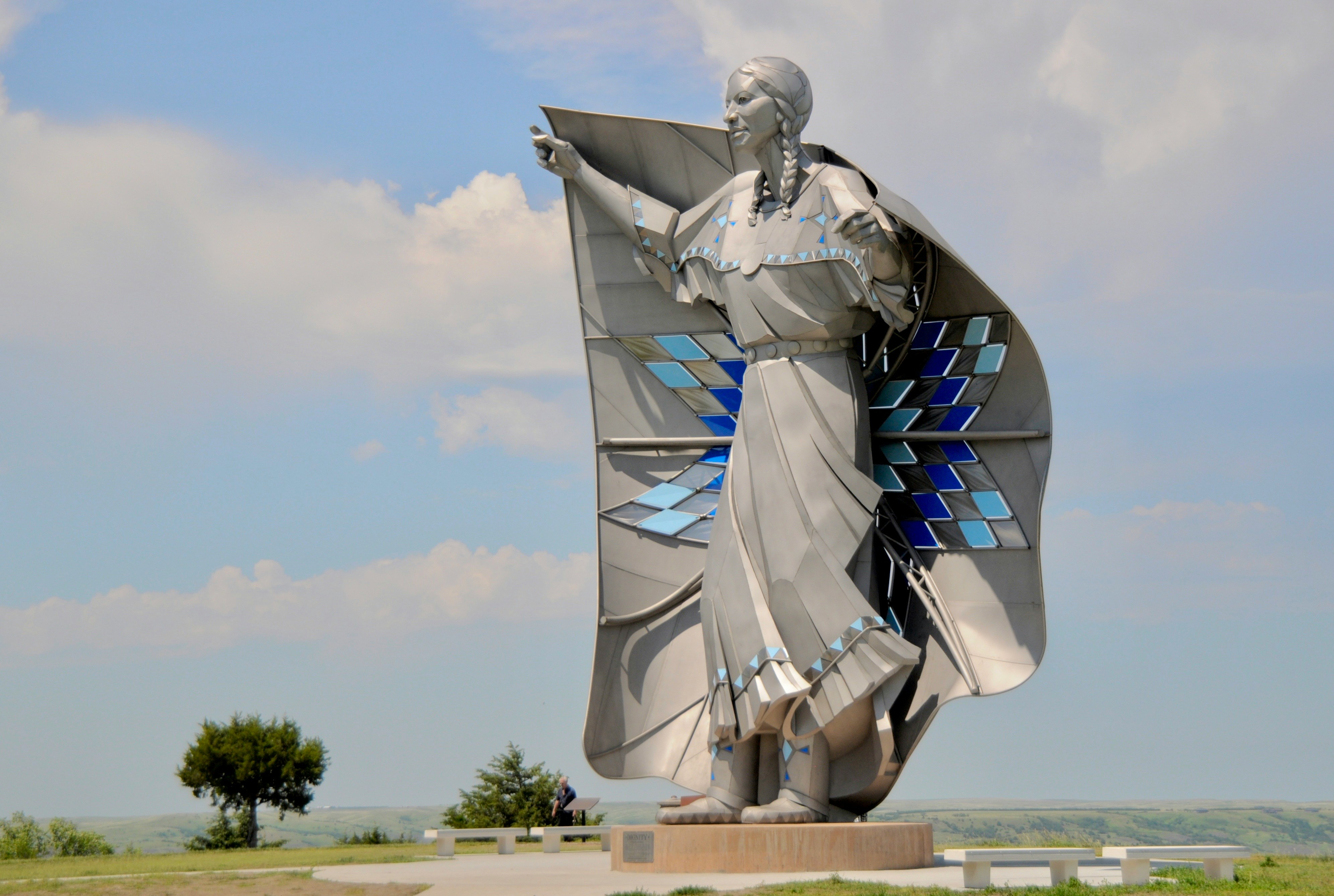 A large stainless-steel statue of a Native woman with long braids, holding out a star quilt inset with blue diamond panels, against an open sky.