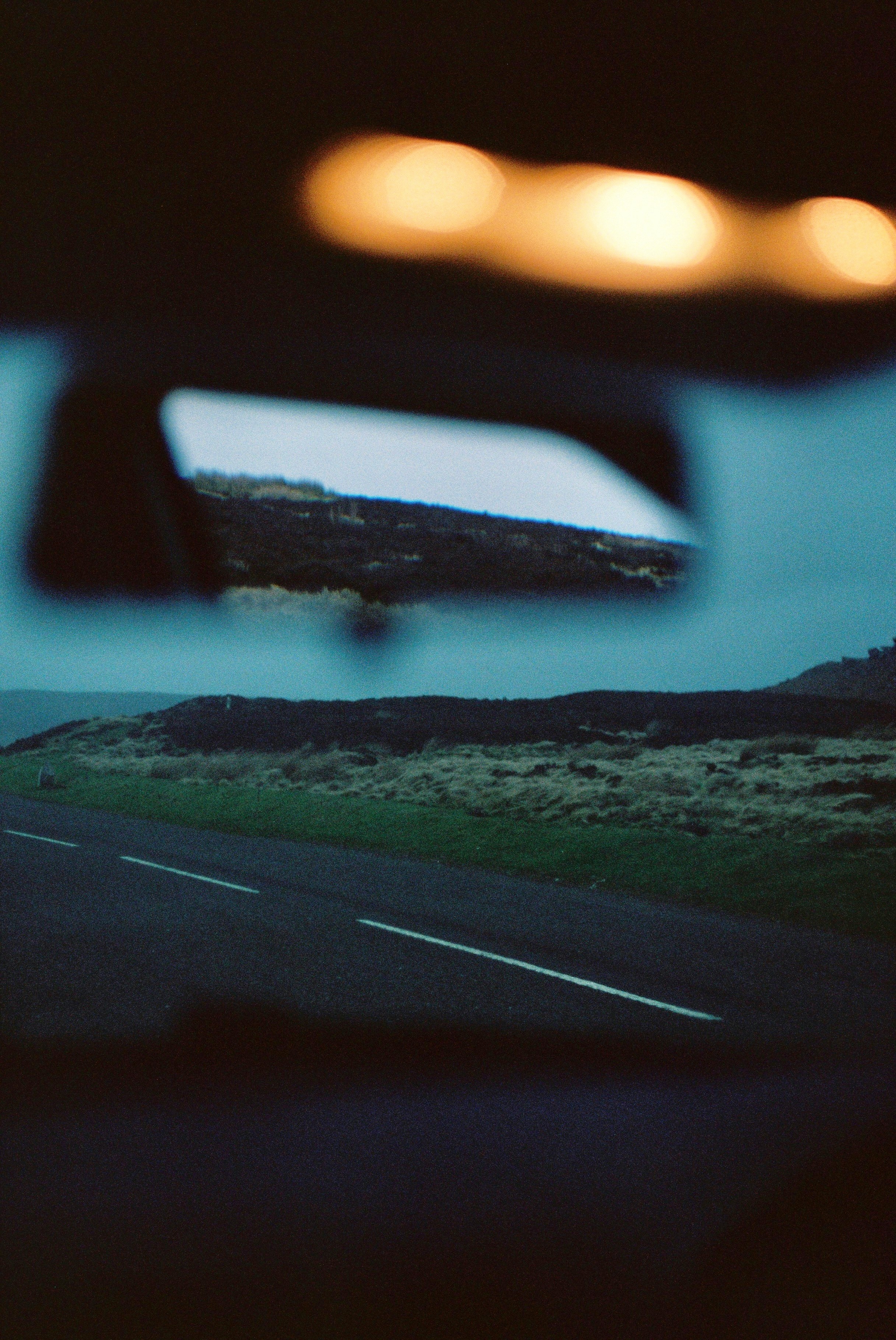 A blurred view through a car's rearview mirror and window, showing a two-lane road, hills, and distant headlights at dusk.