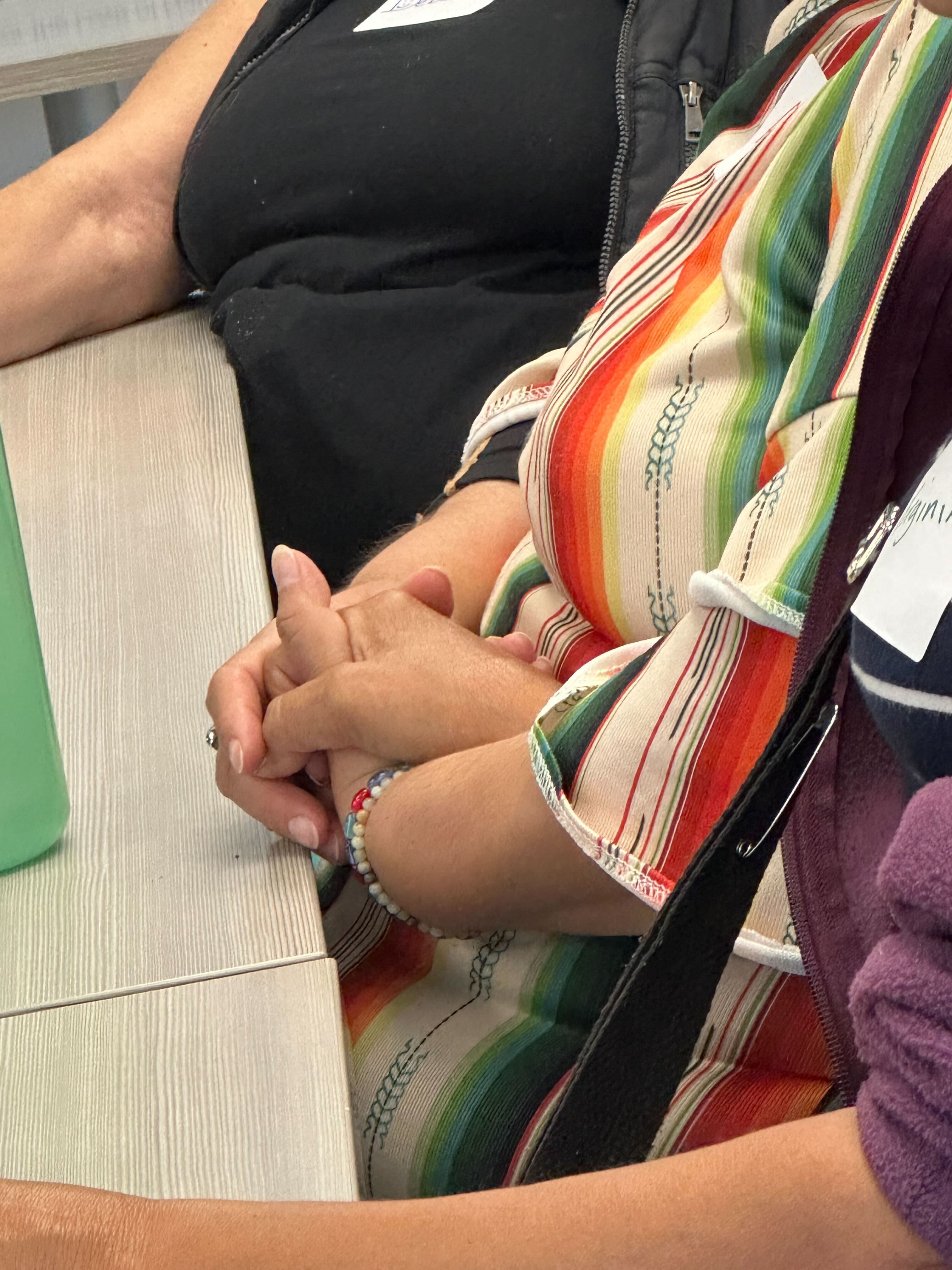 Two people sit close together on a light wood bench, one wearing a brightly striped shirt in red, orange, yellow, and green. In the foreground, a pair of hands rests interlocked in a lap with a beaded bracelet at one wrist.